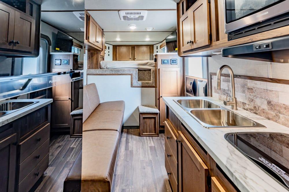 Interior of a modern RV: wood cabinets, double kitchen sink, microwave, and a beige sofa with a raised sleeping area at the back (bed up high).