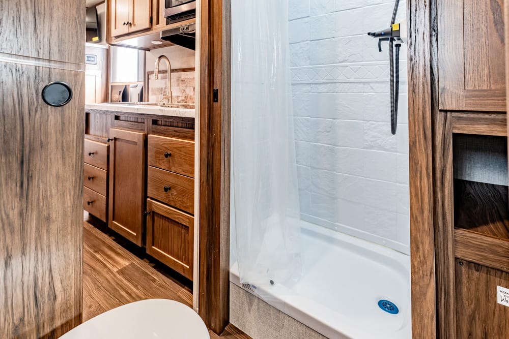 Wood-paneled RV bathroom with a sink and storage on the left, a shower stall with a curtain on the right, and a toilet in the foreground.