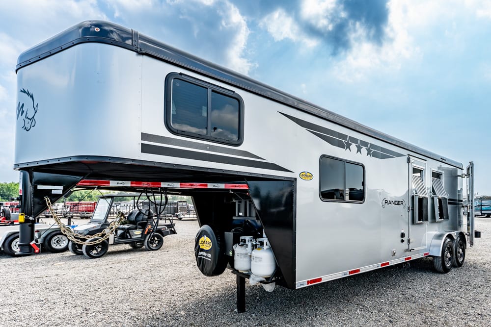 Silver and black fifth-wheel travel trailer with gooseneck hitch parked on gravel, propane tanks visible at front and dual axles on the side side of the trailer visuals and windows visible in daylight.