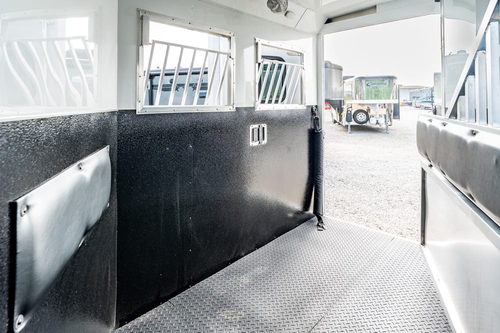 Interior of a horse trailer with black padded walls, a diamond-plate floor, and barred windows; other trailers visible outside through the doorway.",