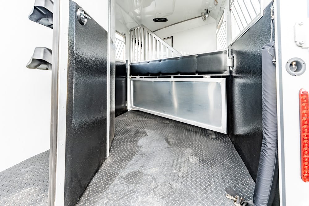 Interior of a horse trailer: black padded divider and metal storage box along the wall, with a diamond‑plate, non-slip floor.