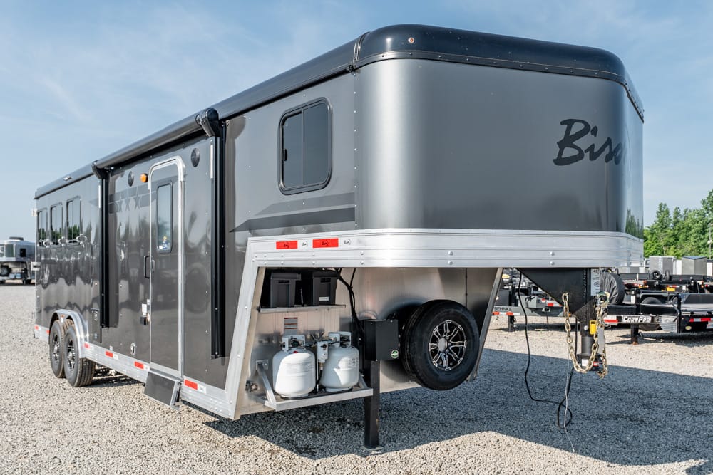 Grey fifth-wheel travel trailer with dual axles, raised front hitch, and exposed propane tanks at the front storage area on a gravel lot