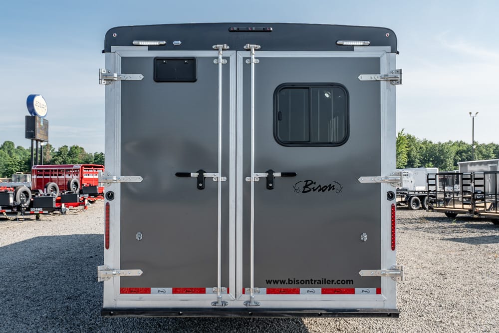 Rear view of a gray Bison horse trailer with double doors, small window, and chrome latches in a trailer lot.