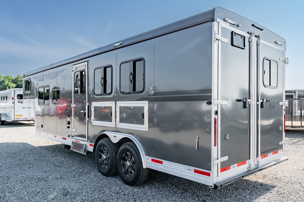 Silver tandem-axle horse trailer parked on a gravel lot, side doors open shows windows and compartments.