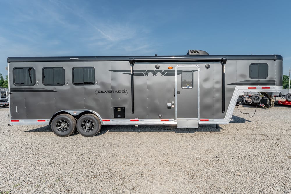 Gray two-axle Silverado travel trailer with side door, windows, and a gooseneck front, parked on gravel under a clear blue sky Glare on metal exterior suggests a new unit.