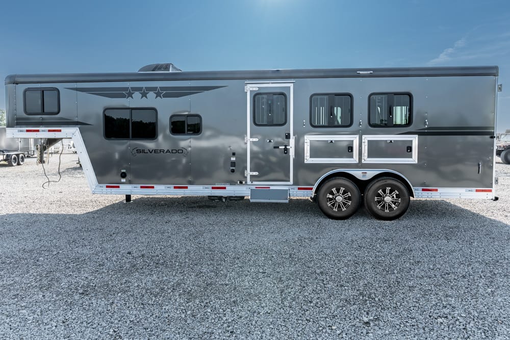 Long silver travel trailer with two axles and side storage compartments, parked on gravel under a blue sky.