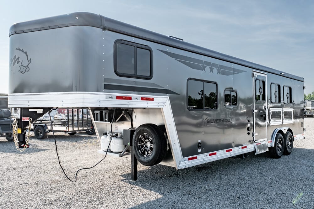 Silver horse trailer with side windows and rear entry doors on a gravel lot, spare tire mounted on the front curb side, propane tank visible undercarriage.