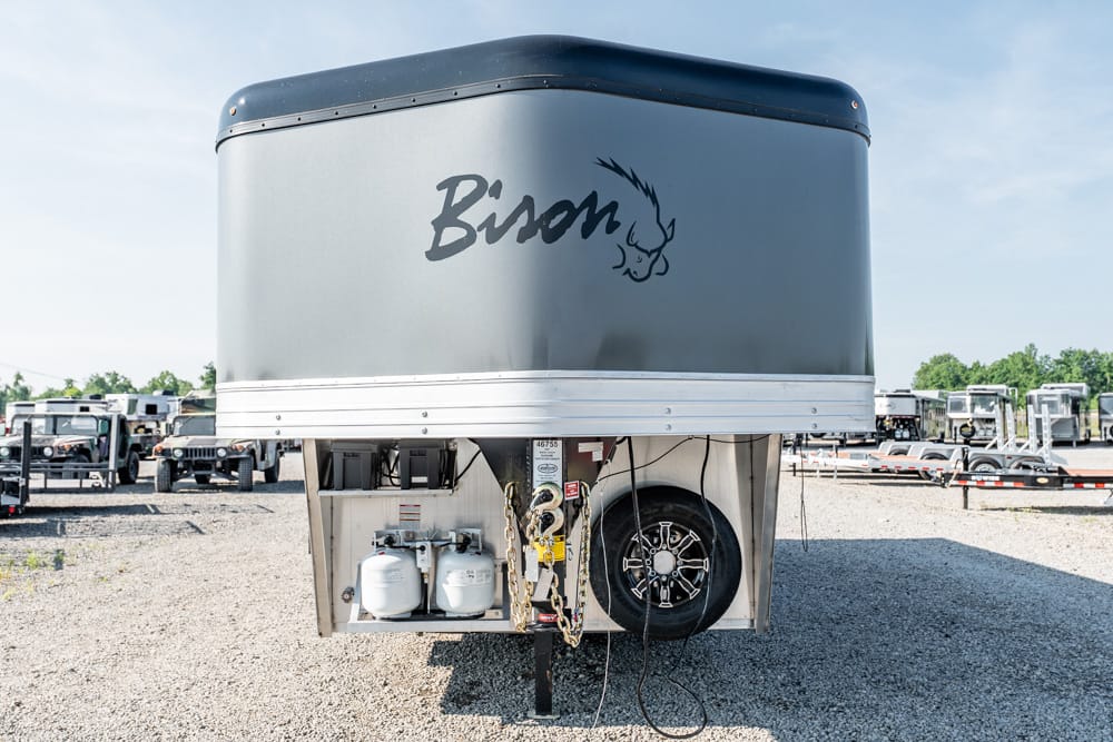 Front view of a gray Bison gooseneck horse trailer with propane tanks, a spare tire, and safety chains at the hitch in a gravel lot.