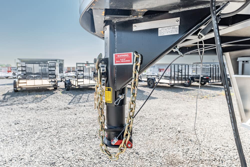 Close-up of a trailer tongue with gold safety chains wrapped around it, red warning label visible on a gravel lot.