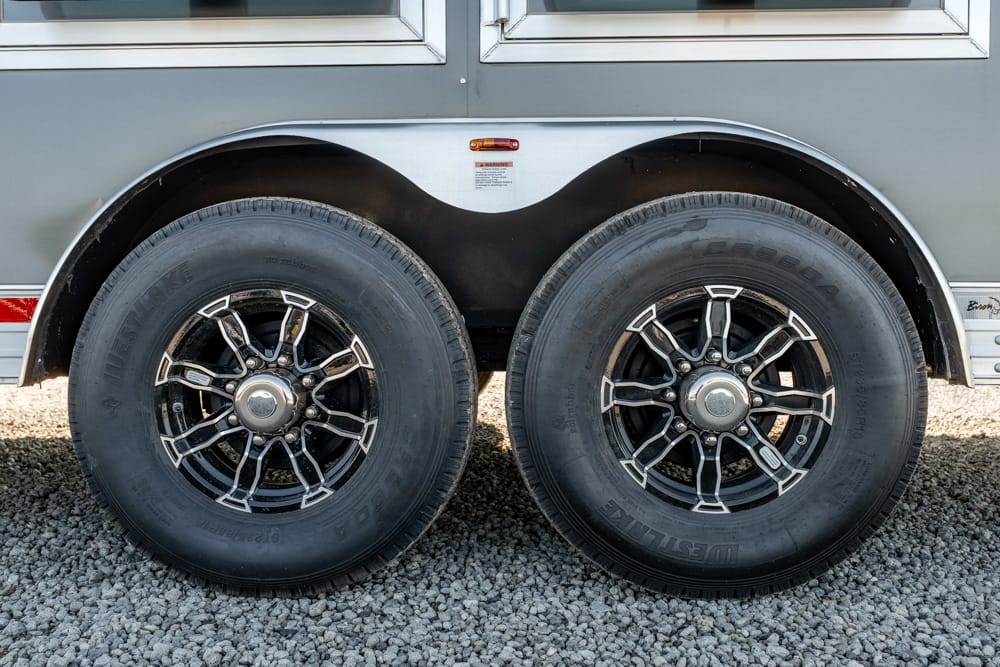 Twin axle wheels on a gray trailer with black and chrome rims, parked on gravel grounds.