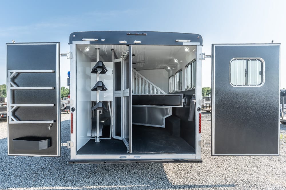 Interior of a horse trailer showing tack storage on the left, vertical saddle racks, a center divider, and a raised breast-height stall area with windows on the right door.