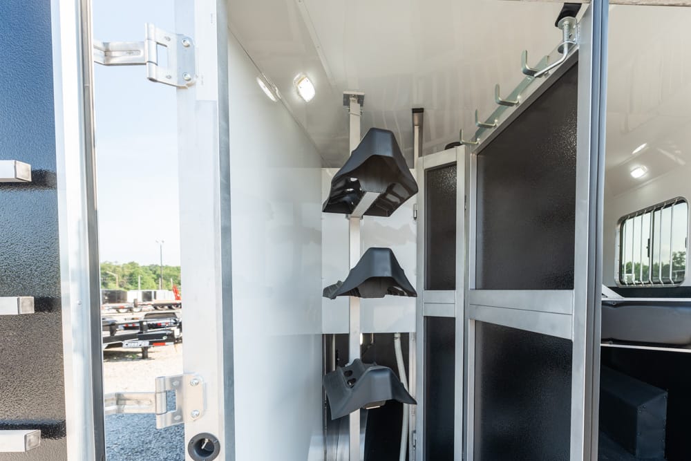 Inside a horse trailer showing a wall-mounted rack with three black saddles hanging on a vertical pole, with metal partitions and hooks along the top edge