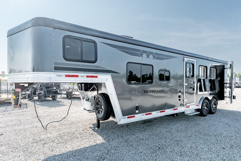 Silverado horse trailer in a gravel lot, grey with windows, ramp doors open at the rear, and a spare tire mounted underneath storage area