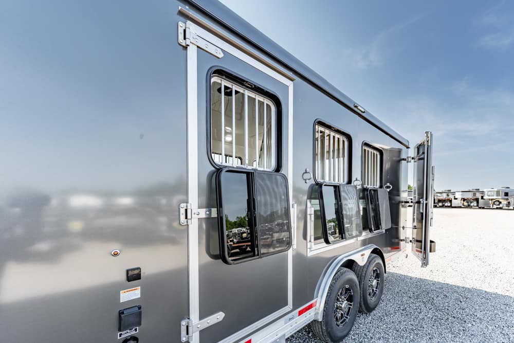 Gray aluminum horse trailer with three barred windows and open side ramp, parked on a gravel lot.