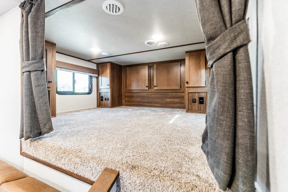 Interior of an RV bedroom with a carpeted raised platform, warm wood cabinets, and a window on the left window light.