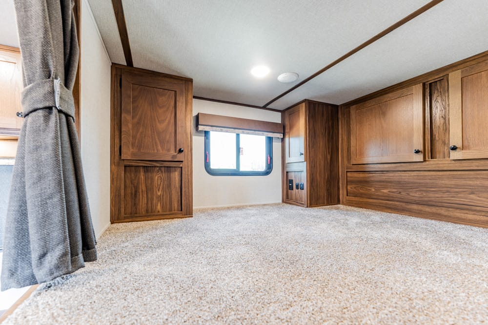 Low-angle view of a cozy RV interior with tall wooden cabinets along the wall, beige carpet, and a small window at center, with gray curtains on the left.