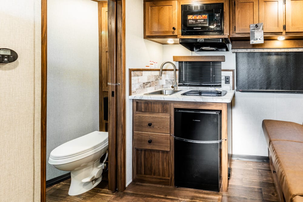 Compact RV kitchenette: sink, stove, microwave above, and a small fridge beneath; bathroom doorway to the left.