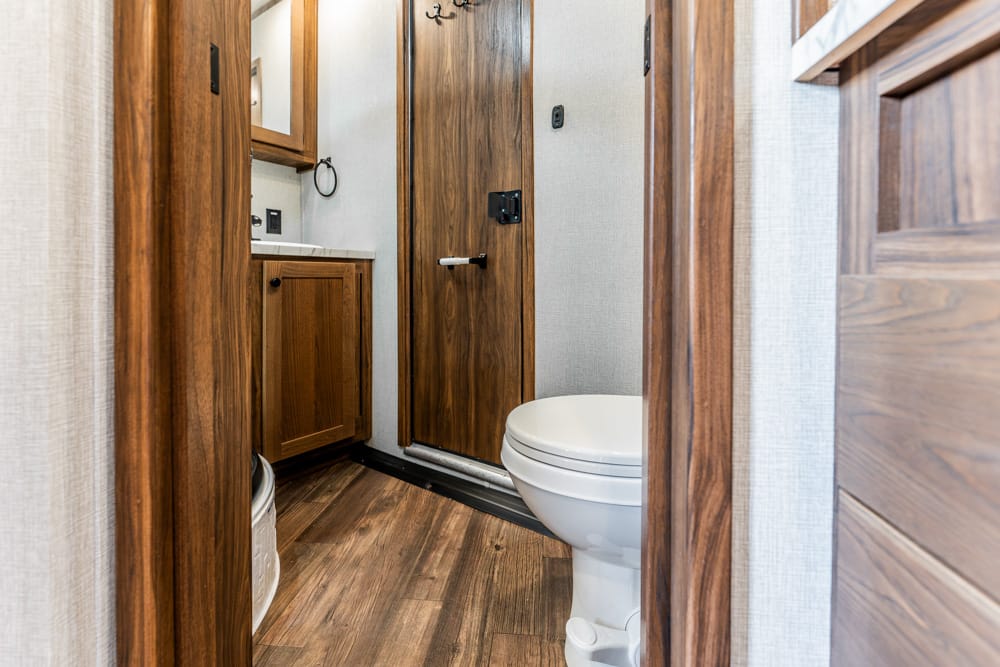Interior of a compact bathroom with a wooden door, vanity, and white toilet on a wood-look floor in a narrow space in a trailer or RV.
