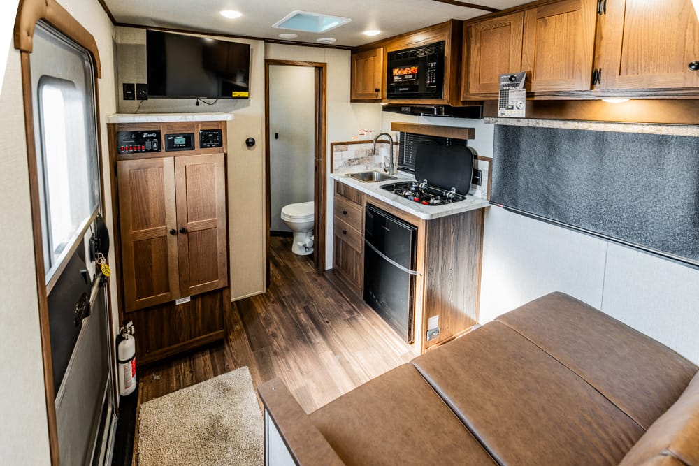 Interior of an RV living area with kitchenette: wood cabinets, microwave, sink, stove, small fridge, and a brown sofa to the right.