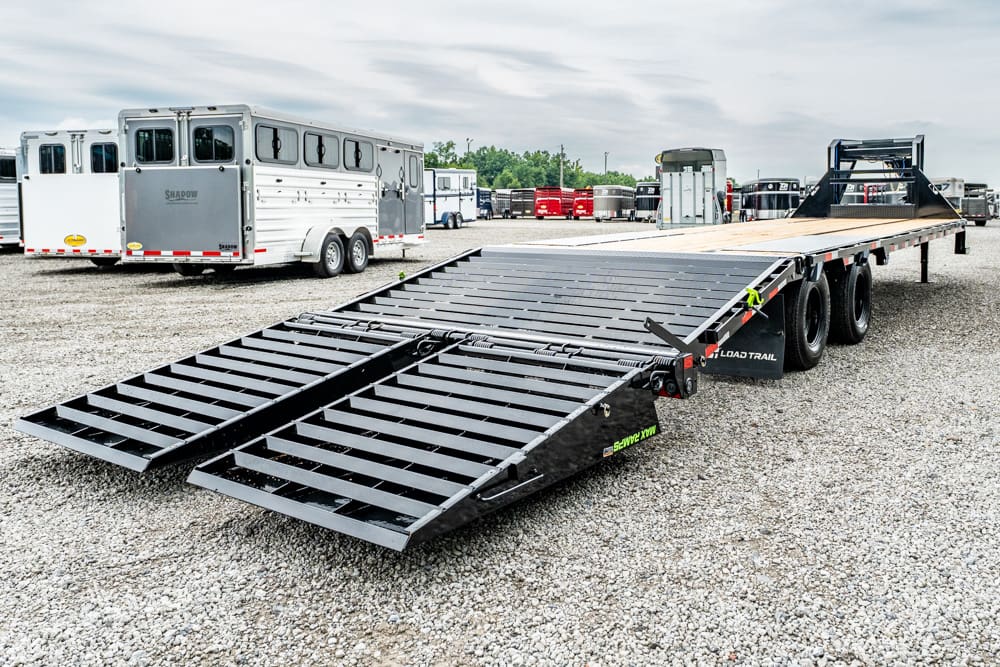 Flatbed trailer with dual ramps extended, parked in a gravel lot with horse trailers in the background; black metal deck, side rails, and tandem axles visible (equipment/ trailer storage yard scene).