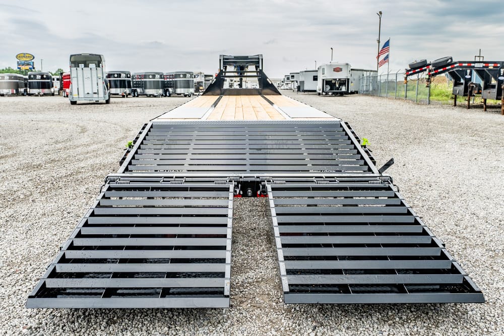Flatbed trailer with two wide metal ramps lowered on a gravel lot beside parked trailers behind it.