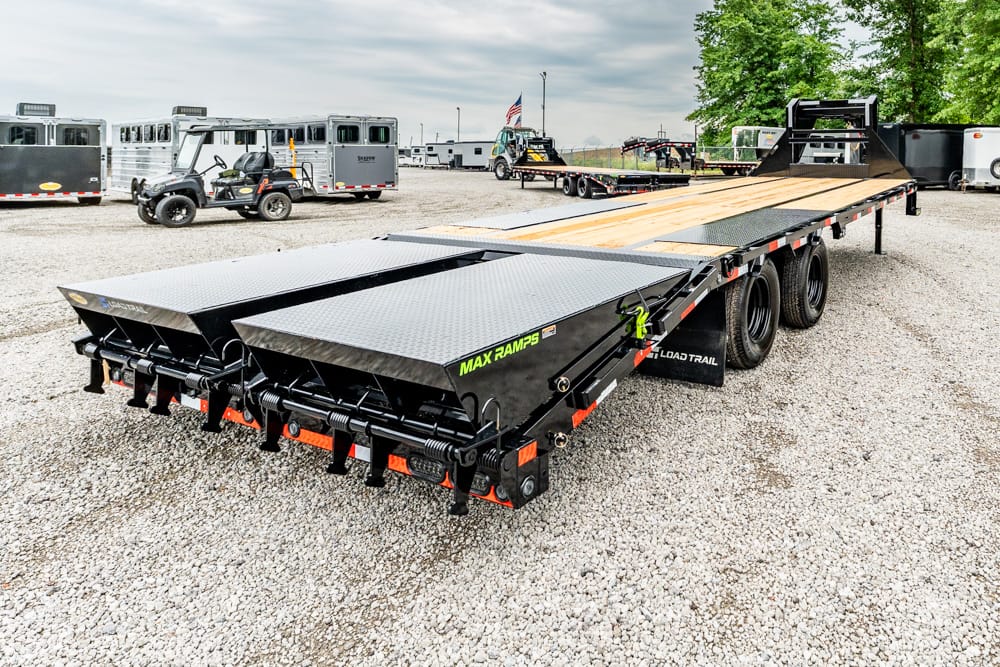 Heavy-duty flatbed trailer with wooden deck and metal ramps on a gravel lot, with other trailers in the background.