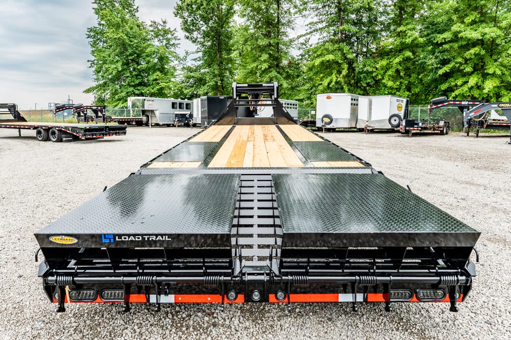 Front view of a Load Trail flatbed trailer with a wooden deck and diamond-plate edges in a gravel trailer yard, trees in the background, ramp up at far end.