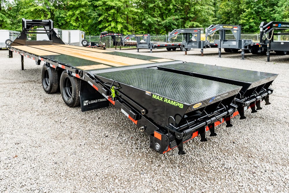 Black Load Trail flatbed trailer with wood deck and fold-out ramps in a gravel lot; other trailers are visible in the background.