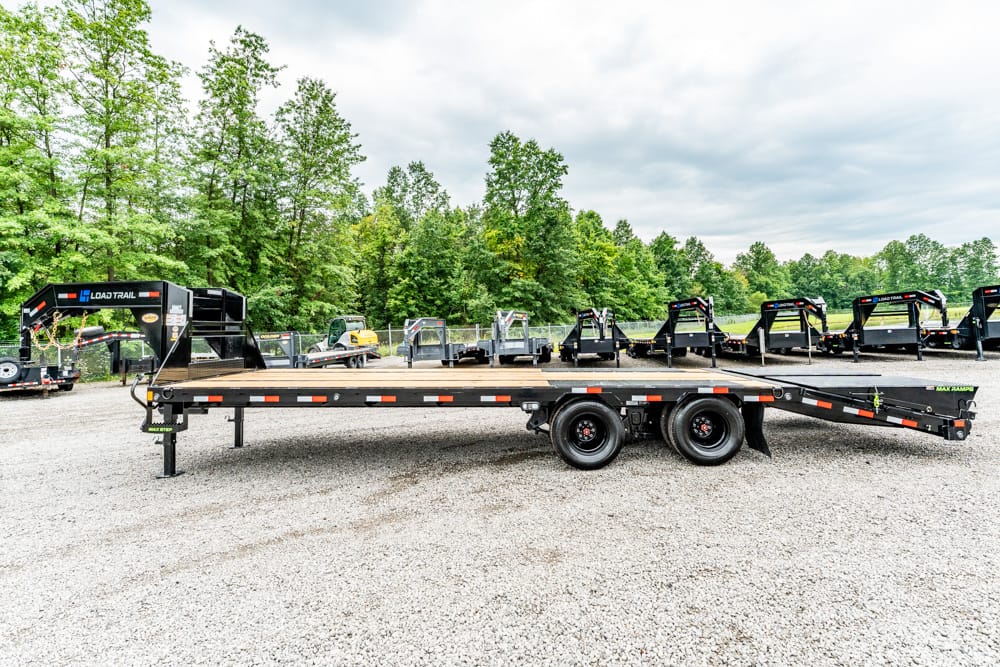 Long black flatbed trailer with two axles parked on a gravel lot, rows of trailers and green trees in the background.