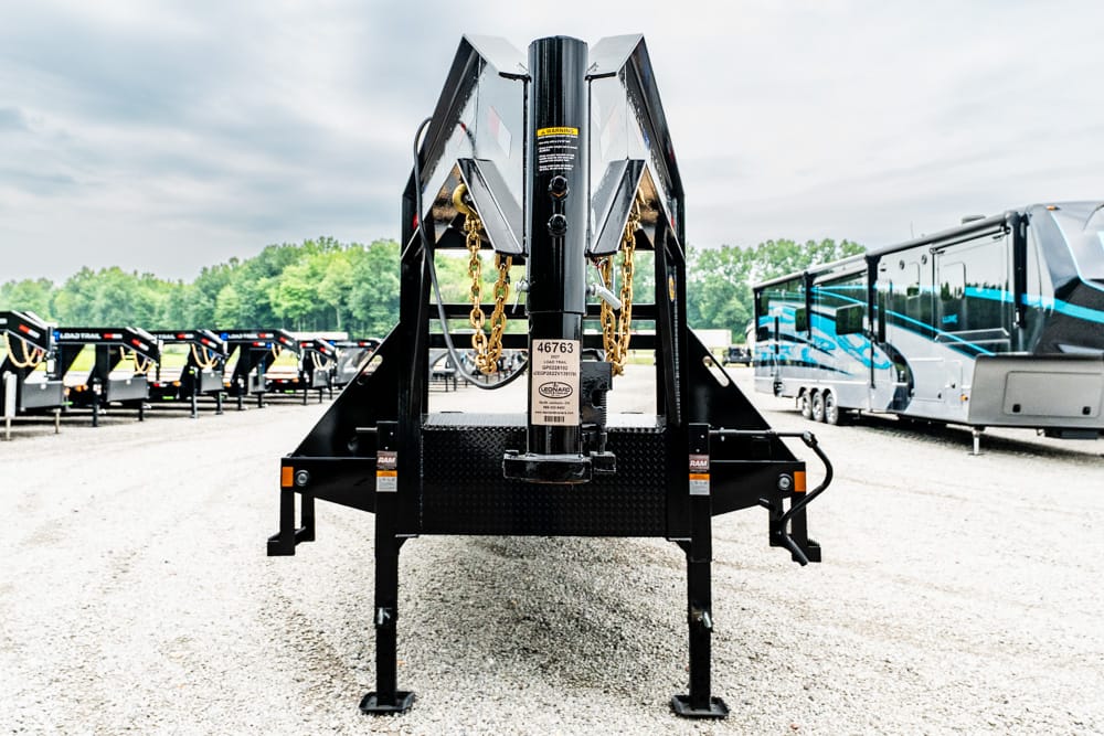 Front view of a black gooseneck trailer hitch with chains and metal framework in a gravel RV lot, surrounded by parked trailers and buses on a cloudy day.
