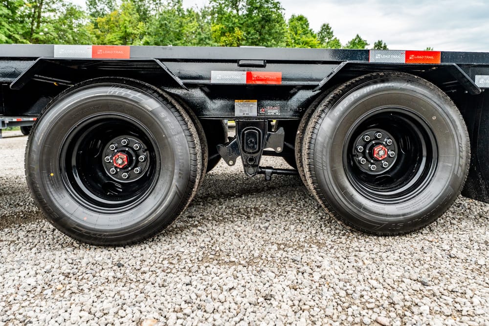 Close-up of a tandem axle on a flatbed trailer, two black wheels with red center caps on a gravel surface.