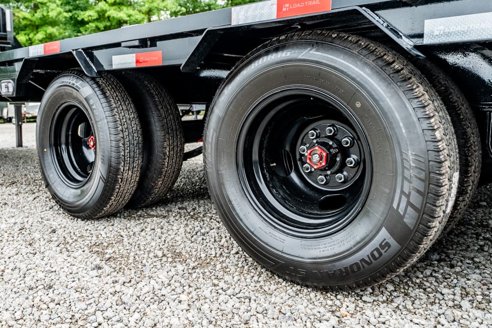 Close-up of dual trailer wheels on a black flatbed trailer parked on a gravel surface with red hub nuts.
