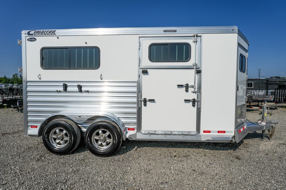 White enclosed horse trailer with two windows, dual rear doors, and tandem wheels on a gravel lot