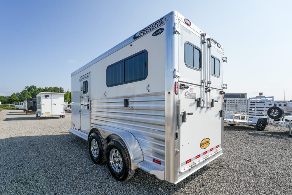 Rear view of a white aluminum horse trailer with double back doors, windows, and tandem wheels on a gravel lot.