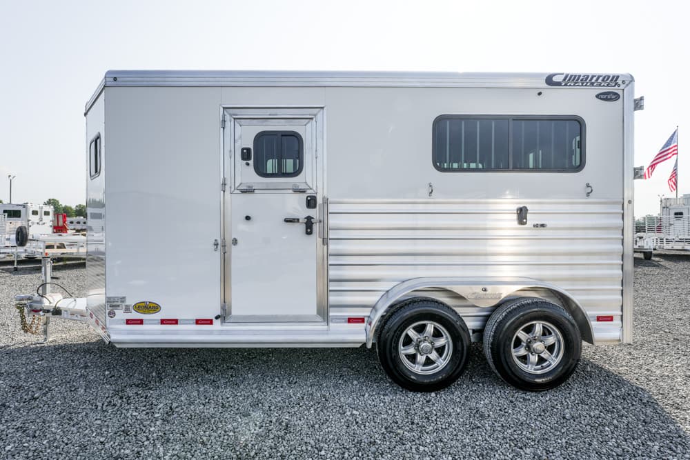 Side view of a silver aluminum horse trailer with a small access door, a large window, and dual rear wheels on gravel ground in a trailer lot.