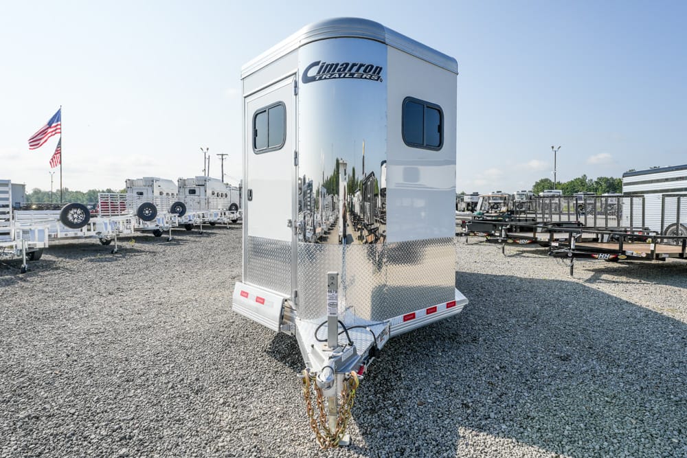 Front view of a shiny silver Cimarron horse trailer with diamond-plate base in a gravel lot, surrounded by other trailers and an American flag nearby.