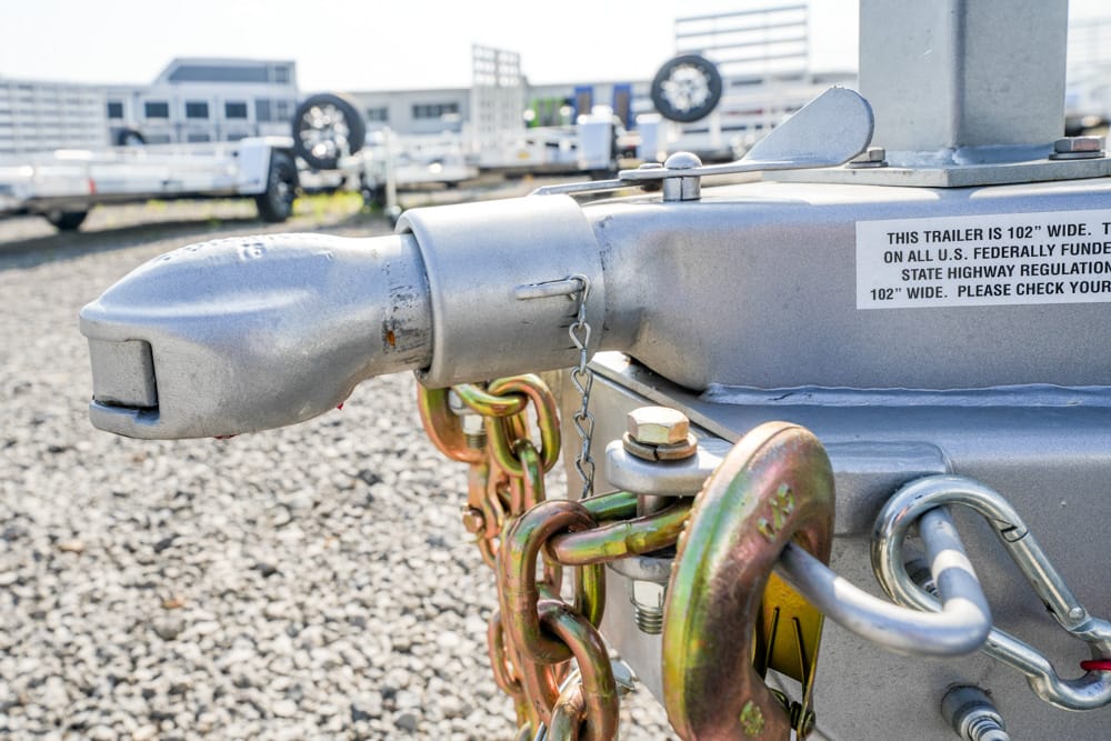 Close-up of a metal trailer hitch with safety chains and a pin, outdoors on a gravel lot.