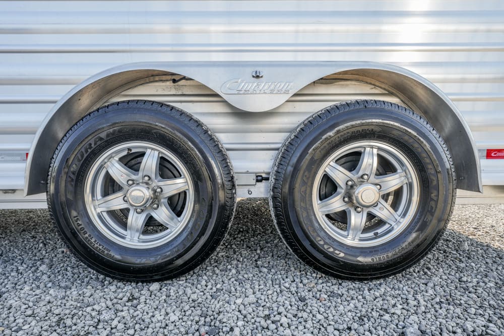 Tandem trailer wheels with silver rims and Goodyear tires, mounted on a metal trailer with curved fenders over a gravel surface.”] ,