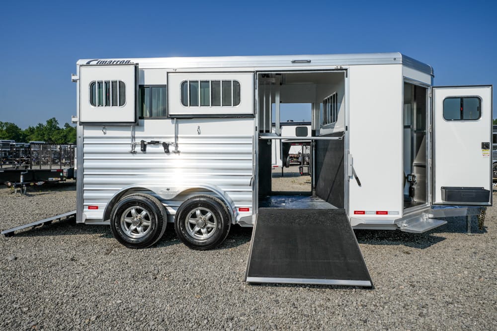 White aluminum horse trailer with a lowered rear ramp on a gravel lot, showing interior stalls and divider panels behind the open door.
