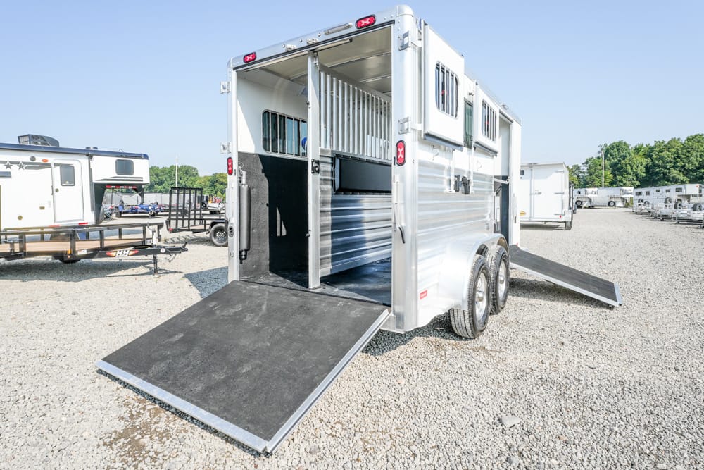 Silver aluminum horse trailer with the rear ramp lowered, showing interior divider stalls on a gravel lot.