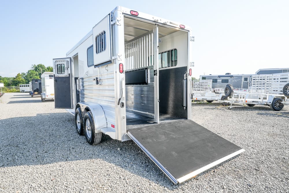 Silver horse trailer with its rear ramp down on a gravel lot, showing the stall divider and windows along the side. Behind it, other trailers are parked in the lot.