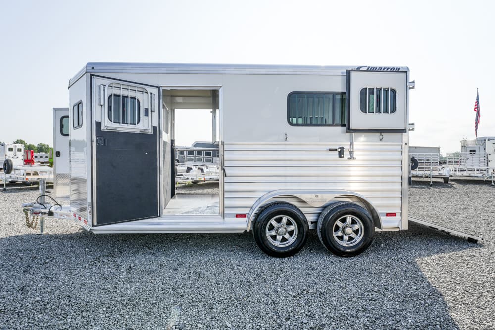 Aluminum horse trailer with two axles on a gravel lot, side door open showing the interior ramp and stall door.