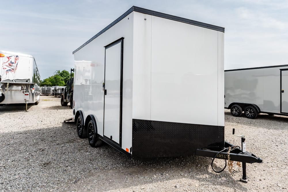 White enclosed cargo trailer with black lower skid plate, parked on gravel lot with other trailers nearby