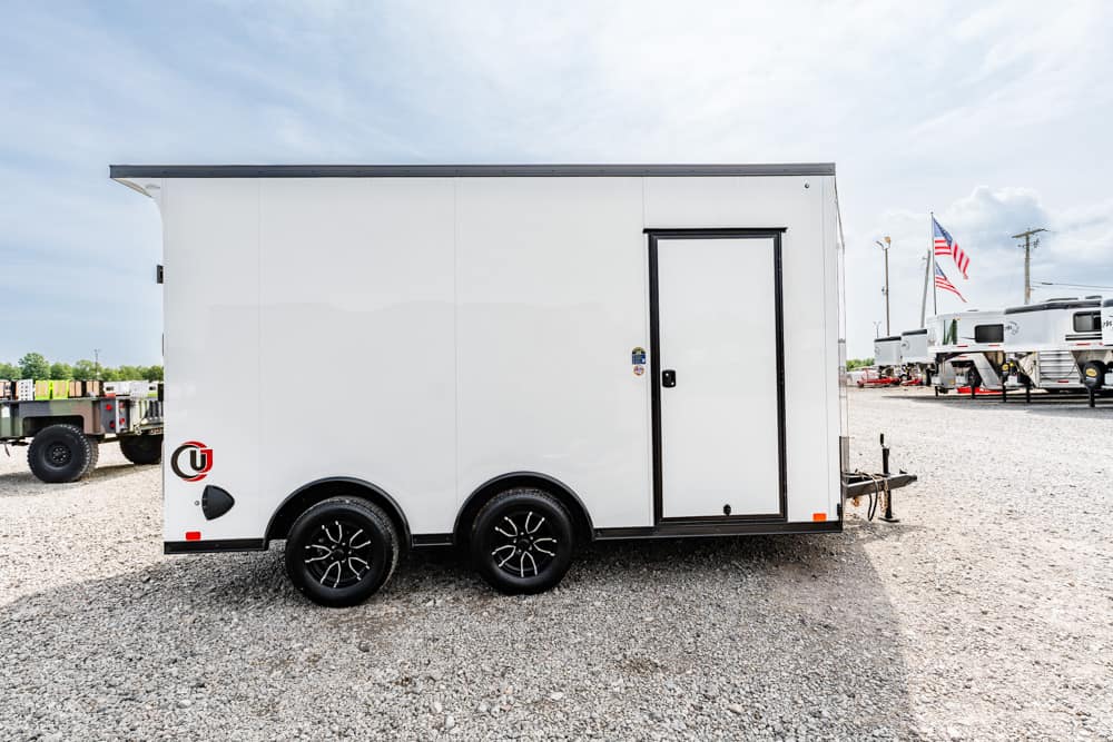 White enclosed cargo trailer with a side door, dual axles, parked on a gravel lot.