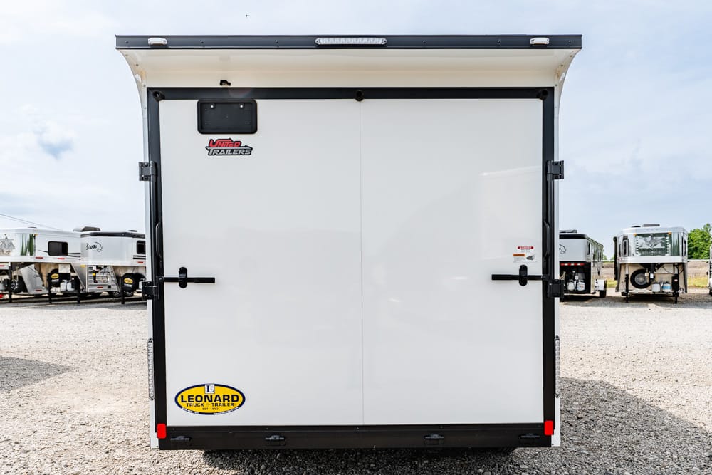 Rear view of a white enclosed cargo trailer with double doors, black latches, and branding stickers in a trailer lot.
