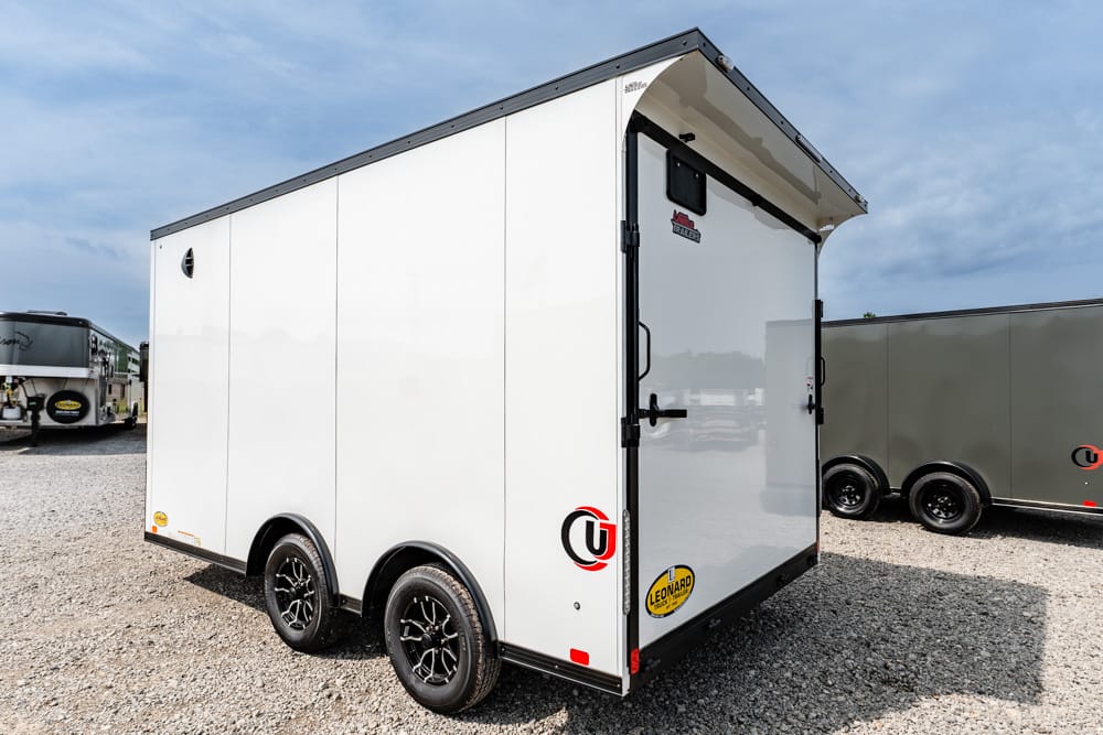 White enclosed cargo trailer with dual axles and black wheels, parked on a gravel lot with other trailers nearby, rear doors closed.