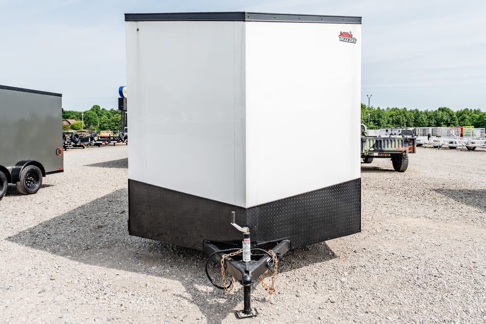 Front view of a white enclosed cargo trailer with a black diamond-plate base on a gravel lot, hitch and safety chains in the foreground.