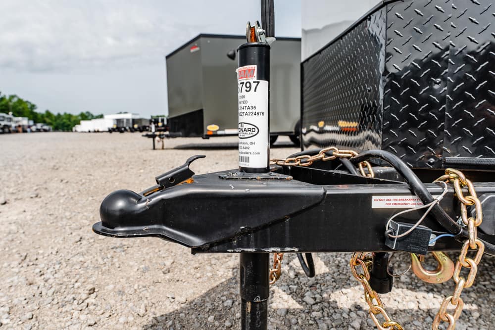 Trailer hitch with a vertical jack labeled '797' and gold safety chains on a gravel lot, with other trailers in the background.