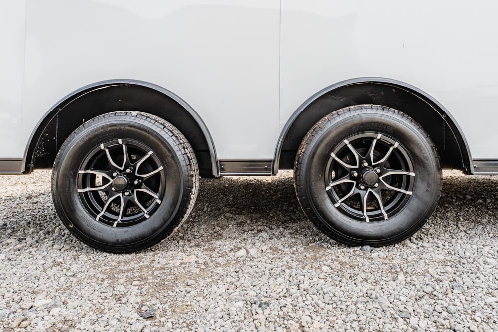 Two black alloy wheels on the side of a light gray trailer, parked on gravel ground.