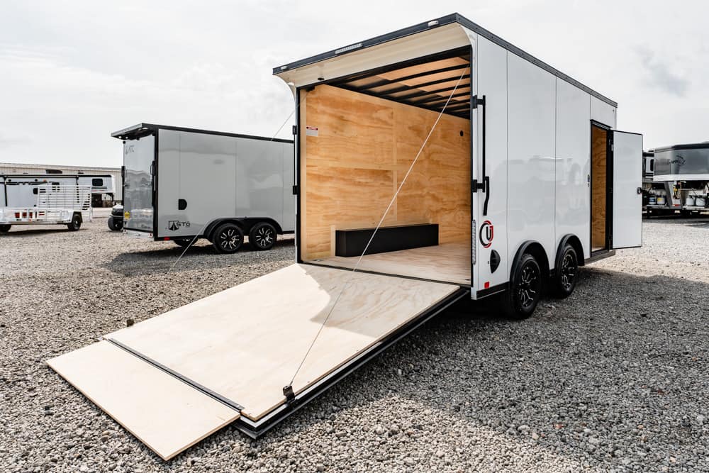 White enclosed cargo trailer with its ramp deployed, showing a plywood interior on a gravel lot.